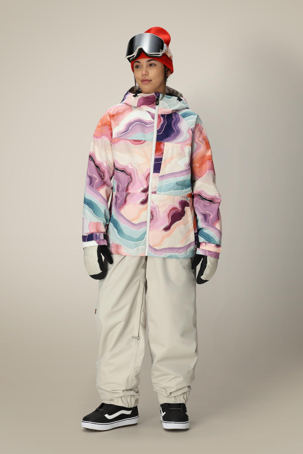 A person wears the 686 Women's Athena™ Insulated Jacket with beige snow pants, white gloves, black sneakers, a red beanie, and ski goggles while standing against a plain background.