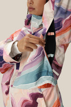 A woman in a 686 Women's Athena™ Insulated Jacket, featuring abstract swirls of pink, purple, blue, and beige, unzips the underarm vent. The waterproof jacket is by 686; her face is partially visible.