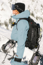 A snowboarder wearing a 686 Women's GORE-TEX Skyline Thermagraph® Jacket stands sideways with a snowboard, black gloves, black goggles, and a black backpack amid snowy mountains and trees.