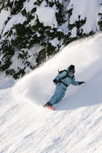 A snowboarder wears the 686 Women's GORE-TEX Skyline Thermagraph® Jacket, gliding down a snowy slope and kicking up powder, with snow-covered evergreens in the background.