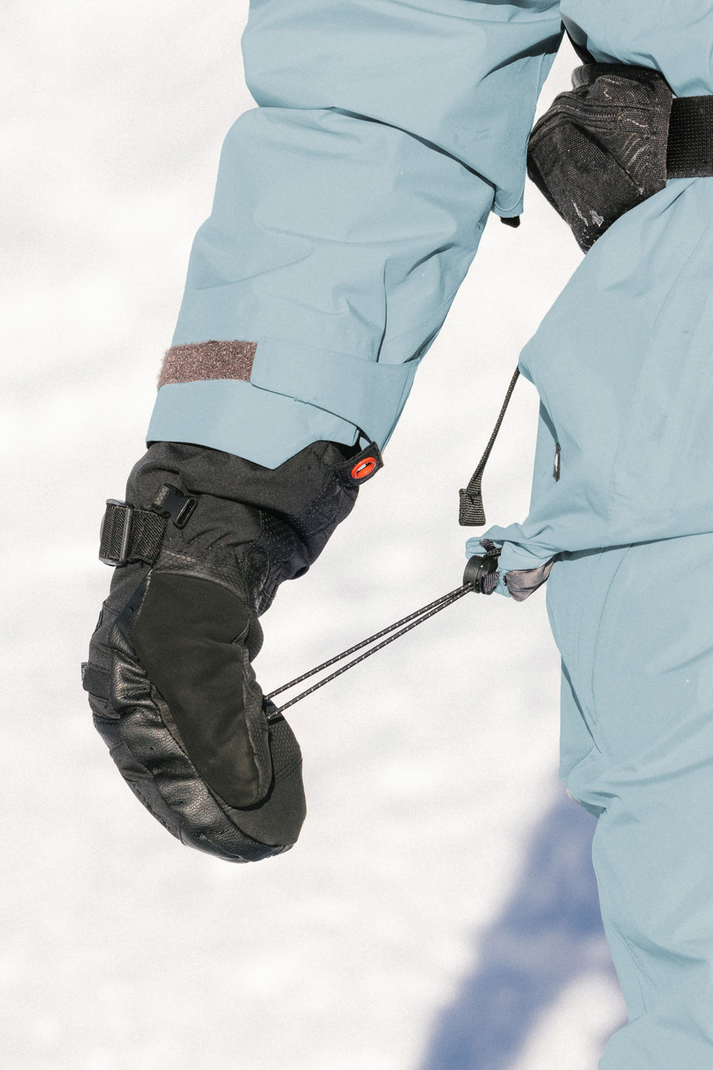 A person wearing the 686 Women's GORE-TEX Skyline Thermagraph® Jacket in light blue and black insulated gloves adjusts the waist drawstring, with snow visible in the background.