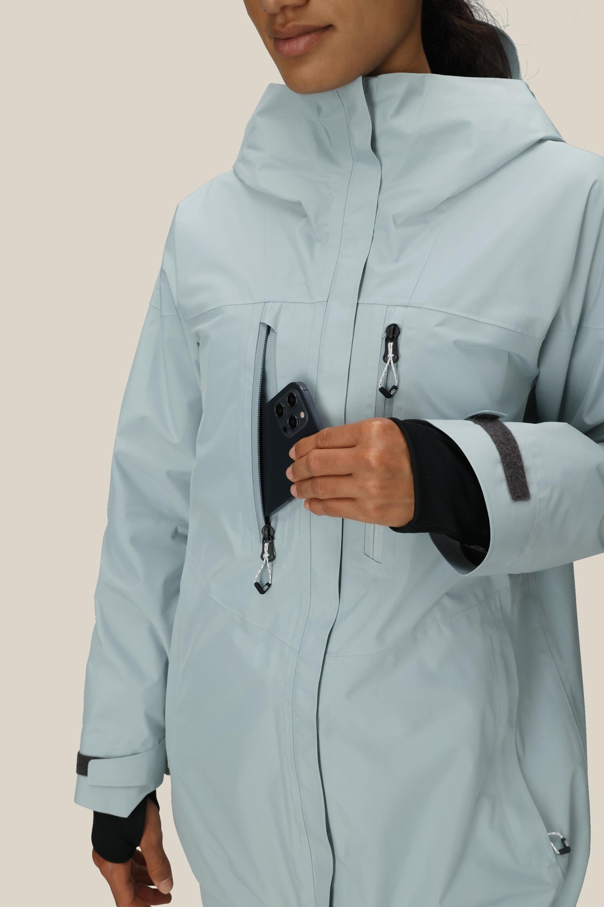 A woman in a light blue 686 Women's GORE-TEX Skyline Thermagraph® Jacket places a smartphone into its zippered chest pocket against a plain, neutral background.