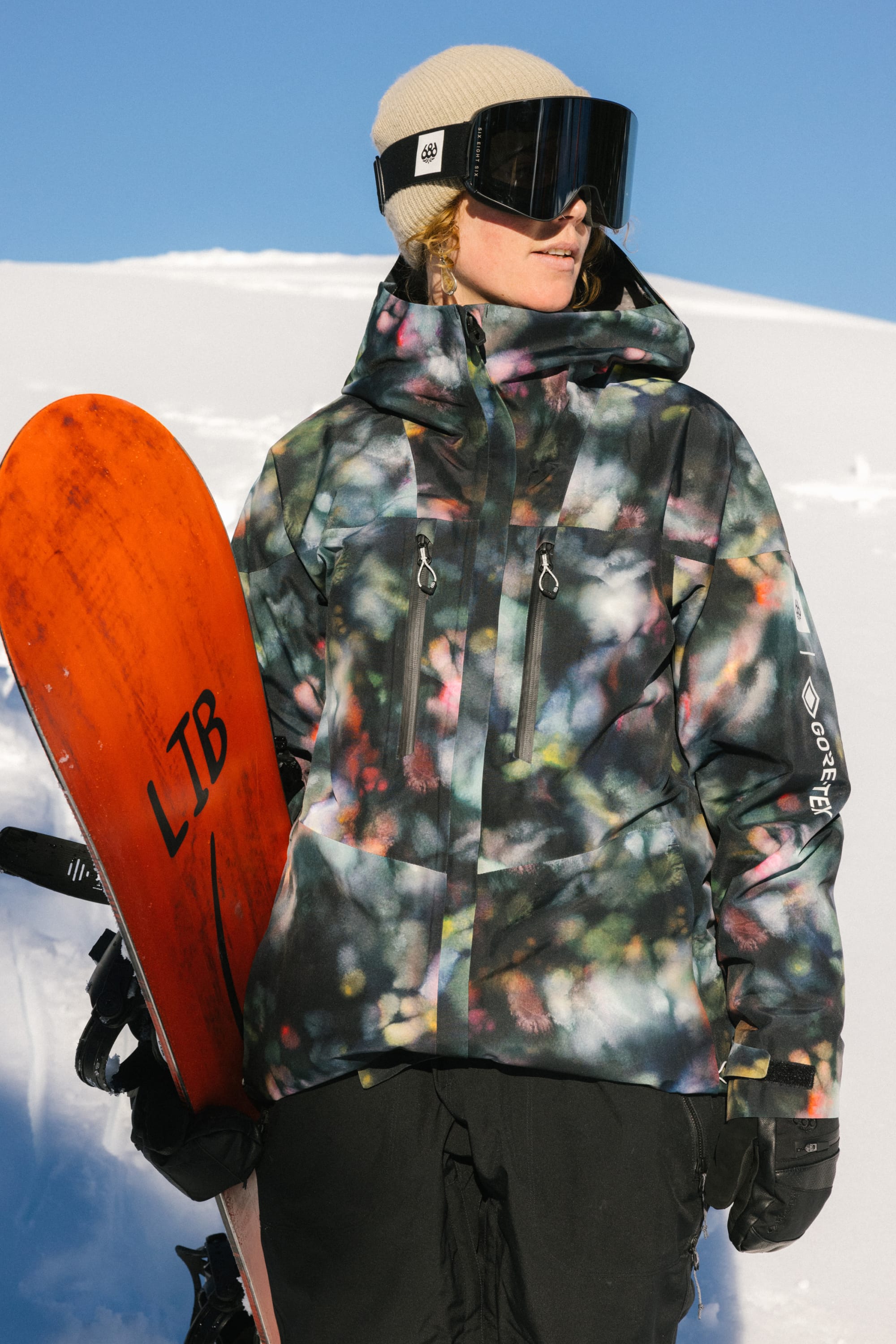 Wearing a 686 Women's GORE-TEX Skyline Thermagraph® Jacket with a bold pattern, black pants, tan beanie, and large ski goggles, a woman holds an orange snowboard in the sunny, snowy outdoors.