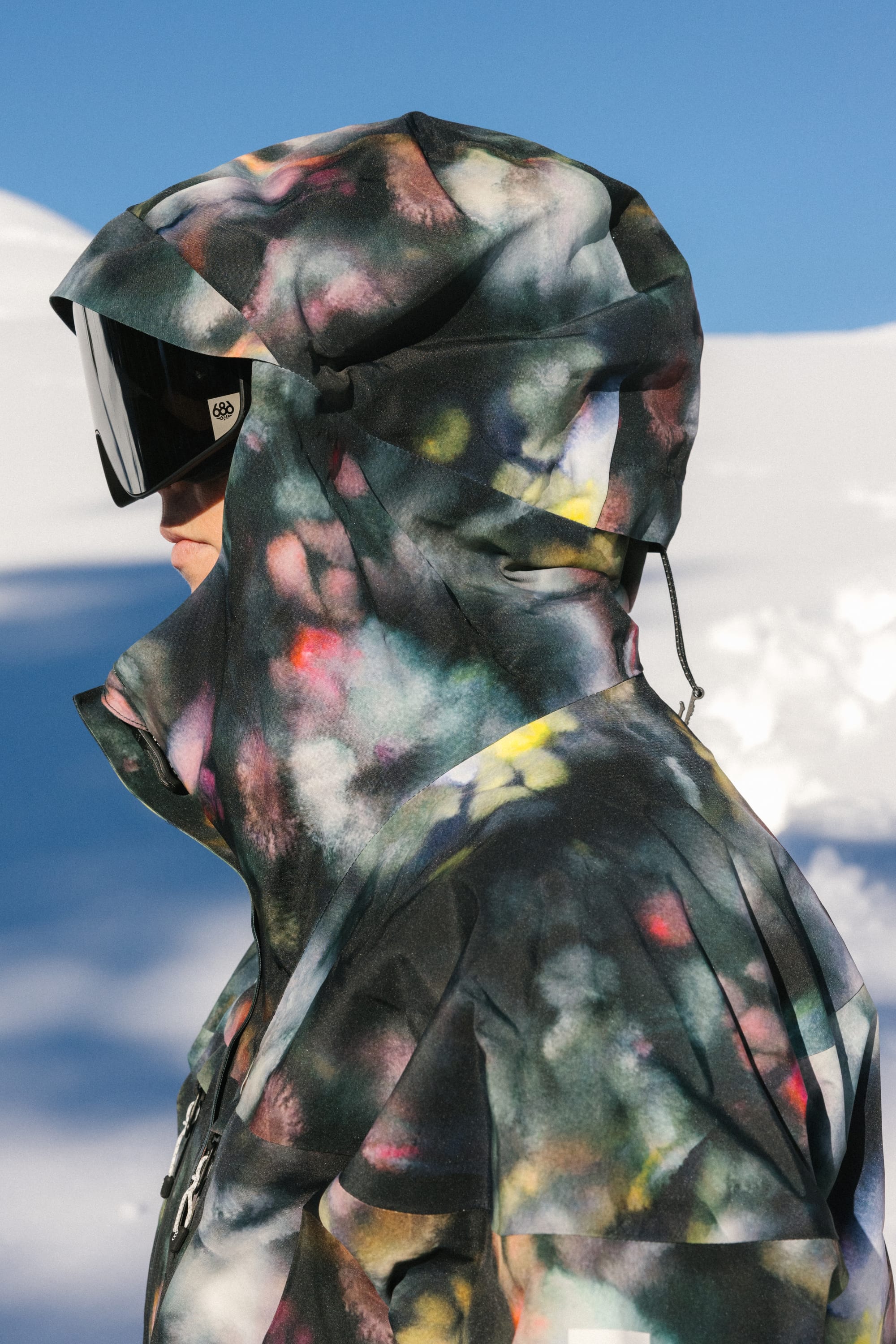 A woman wearing the 686 Women's GORE-TEX Skyline Thermagraph® Jacket and reflective ski goggles stands in profile against a snowy outdoor background.