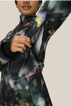 A woman unzips the side vent of a 686 Women's GORE-TEX Skyline Thermagraph® Jacket, revealing an inner layer. The image is cropped to show just her lower face and upper body against a plain background.
