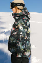 A person wearing the 686 Women's GORE-TEX Skyline Thermagraph® Jacket and ski goggles stands in profile on a snowy landscape under a clear blue sky.