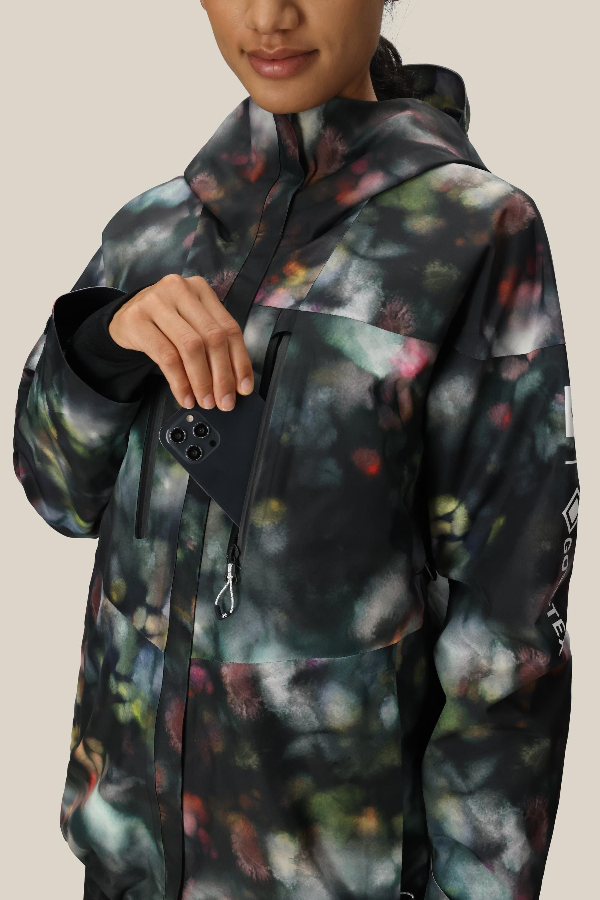 A person wears the 686 Women's GORE-TEX Skyline Thermagraph® Jacket, featuring an abstract pattern in green, pink, and white on a dark background. The high-collar design has a chest pocket where a black smartphone is partially tucked. Brand: 686.