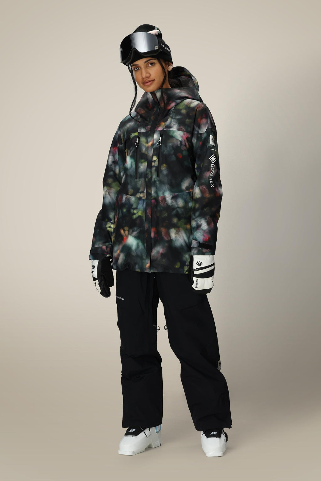 A person wearing the 686 Women's GORE-TEX Skyline Thermagraph® Jacket stands facing forward, paired with black snow pants, white ski gloves, black ski goggles, and white ski boots against a plain background.
