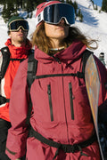 Two snowboarders in red 686 Women's GORE-TEX Skyline Thermagraph® Jackets, with helmets and goggles, stand on a snowy mountain. The front snowboarder looks up holding a board, the other faces forward. Pine trees and bright sunlight are behind them.