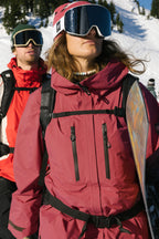 Two snowboarders in red 686 Women's GORE-TEX Skyline Thermagraph® Jackets, with helmets and goggles, stand on a snowy mountain. The front snowboarder looks up holding a board, the other faces forward. Pine trees and bright sunlight are behind them.
