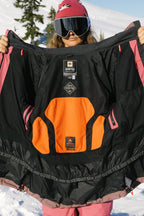 Wearing pink ski gear and goggles, a person displays the detailed lining of the 686 Women's GORE-TEX Skyline Thermagraph® Jacket in black and orange, with snowy mountains and trees in the background.