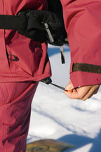 A person adjusts the hem drawstring of their 686 Women's GORE-TEX Skyline Thermagraph® Jacket, standing on snow. Only their hand and part of the maroon jacket and pants are visible.