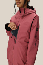 A woman in a 686 Women's GORE-TEX Skyline Thermagraph® Jacket places a black smartphone into the chest pocket. Only the lower part of her face is visible, with a plain beige background.