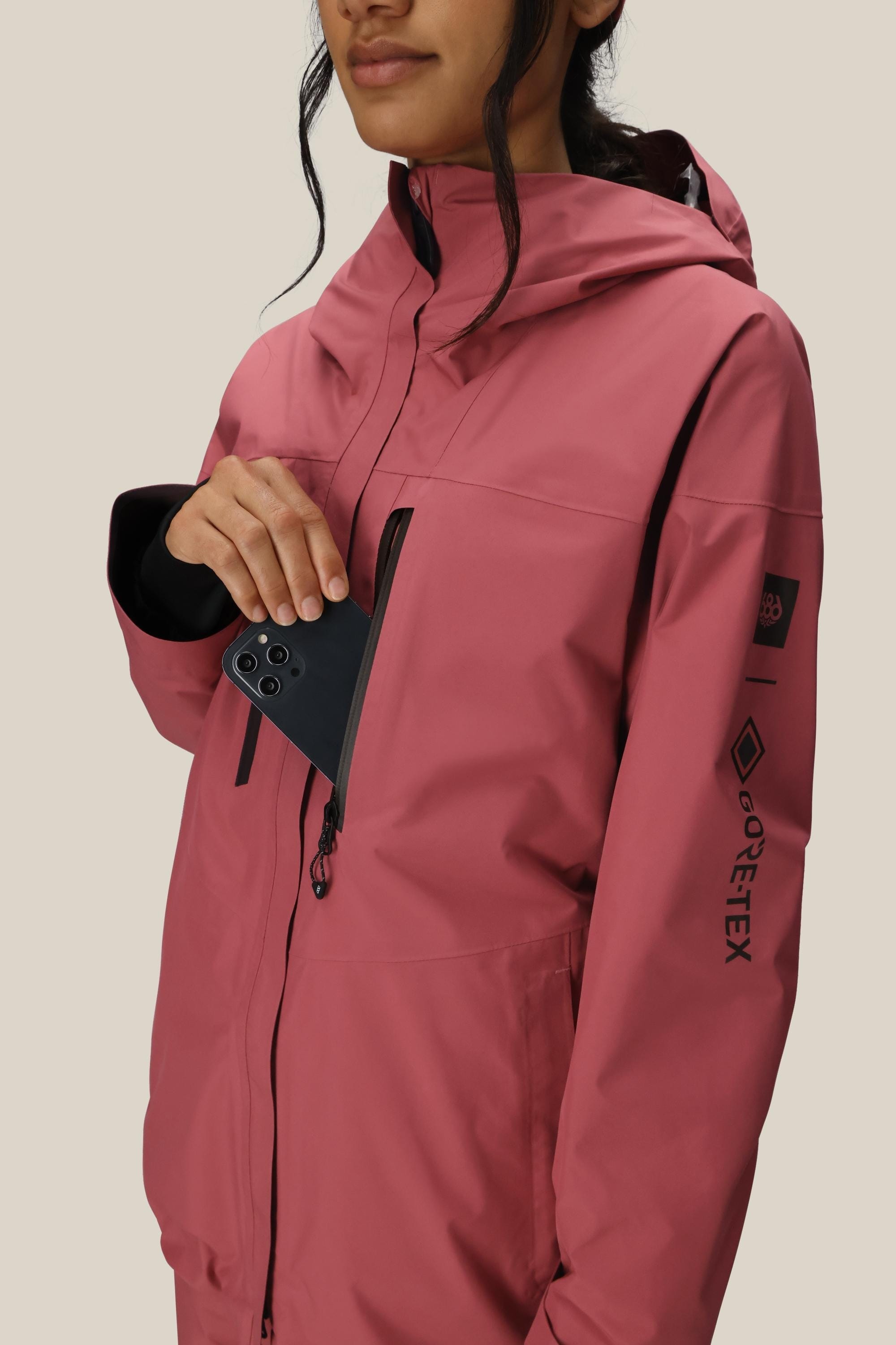 A woman in a 686 Women's GORE-TEX Skyline Thermagraph® Jacket places a black smartphone into the chest pocket. Only the lower part of her face is visible, with a plain beige background.