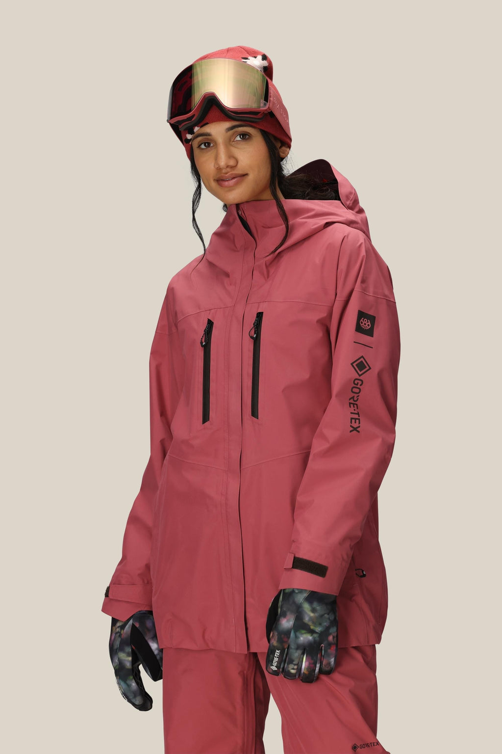 A woman wears the 686 Women's GORE-TEX Skyline Thermagraph® Jacket in rose pink with black zippers and GORE-TEX logo, paired with matching pants, patterned gloves, a helmet, and mirrored ski goggles against a neutral background.