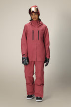 A person wears a 686 Women's GORE-TEX Skyline Thermagraph® Jacket in pink with matching pants, black sneakers, patterned gloves, and a red helmet with reflective goggles, standing against a neutral background.