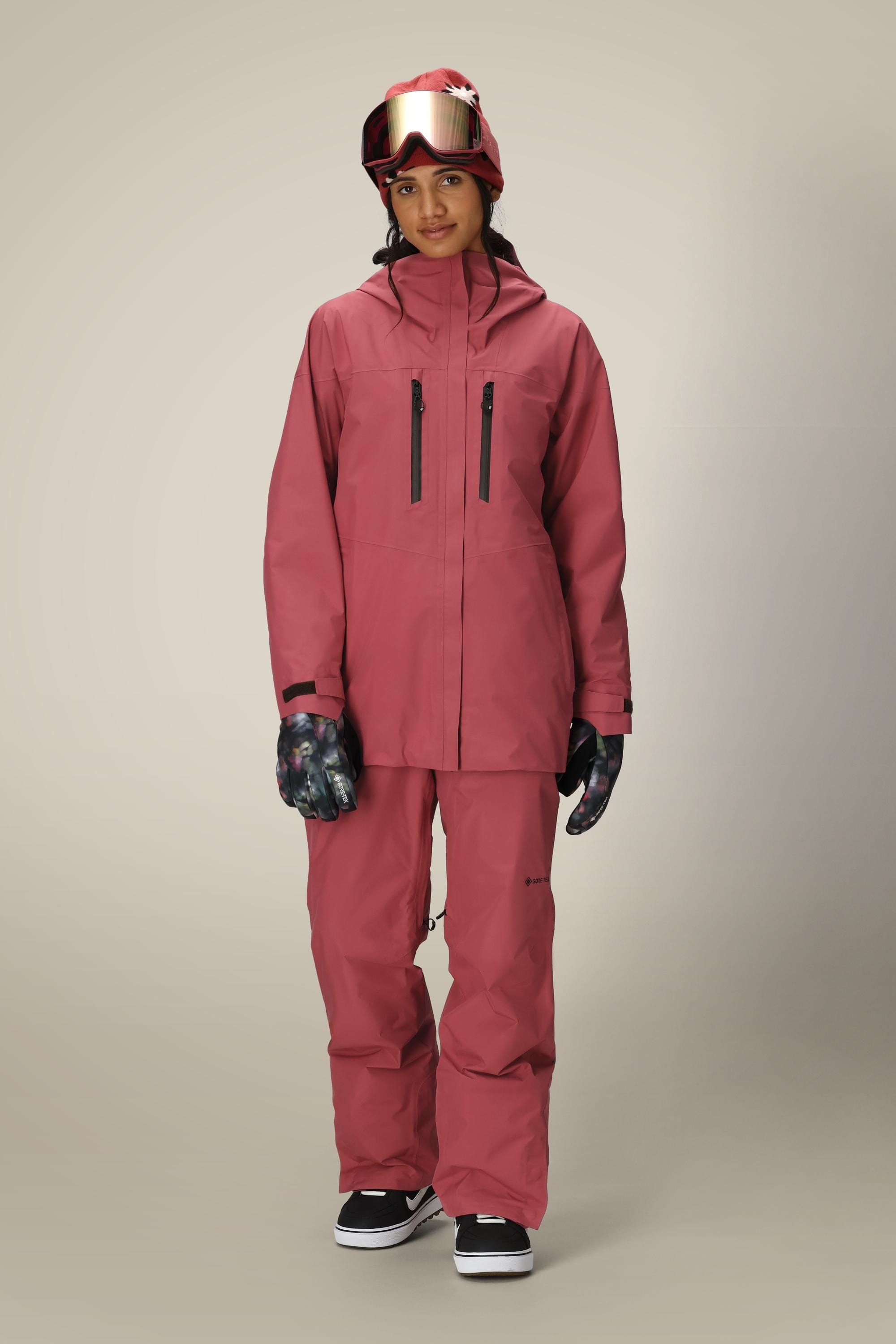 A person wears a 686 Women's GORE-TEX Skyline Thermagraph® Jacket in pink with matching pants, black sneakers, patterned gloves, and a red helmet with reflective goggles, standing against a neutral background.