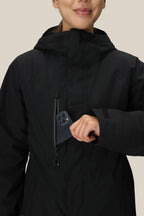 A woman wearing the 686 Women's GORE-TEX Willow™ Insulated Jacket smiles as she places her smartphone into the jacket’s zippered chest pocket.