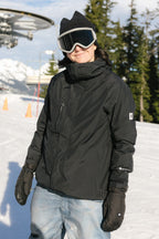Wearing a 686 Women's GORE-TEX Willow™ Insulated Jacket, ski gloves, goggles, and a beanie, a person stands on a snowy slope with trees and ski lift equipment in the background.