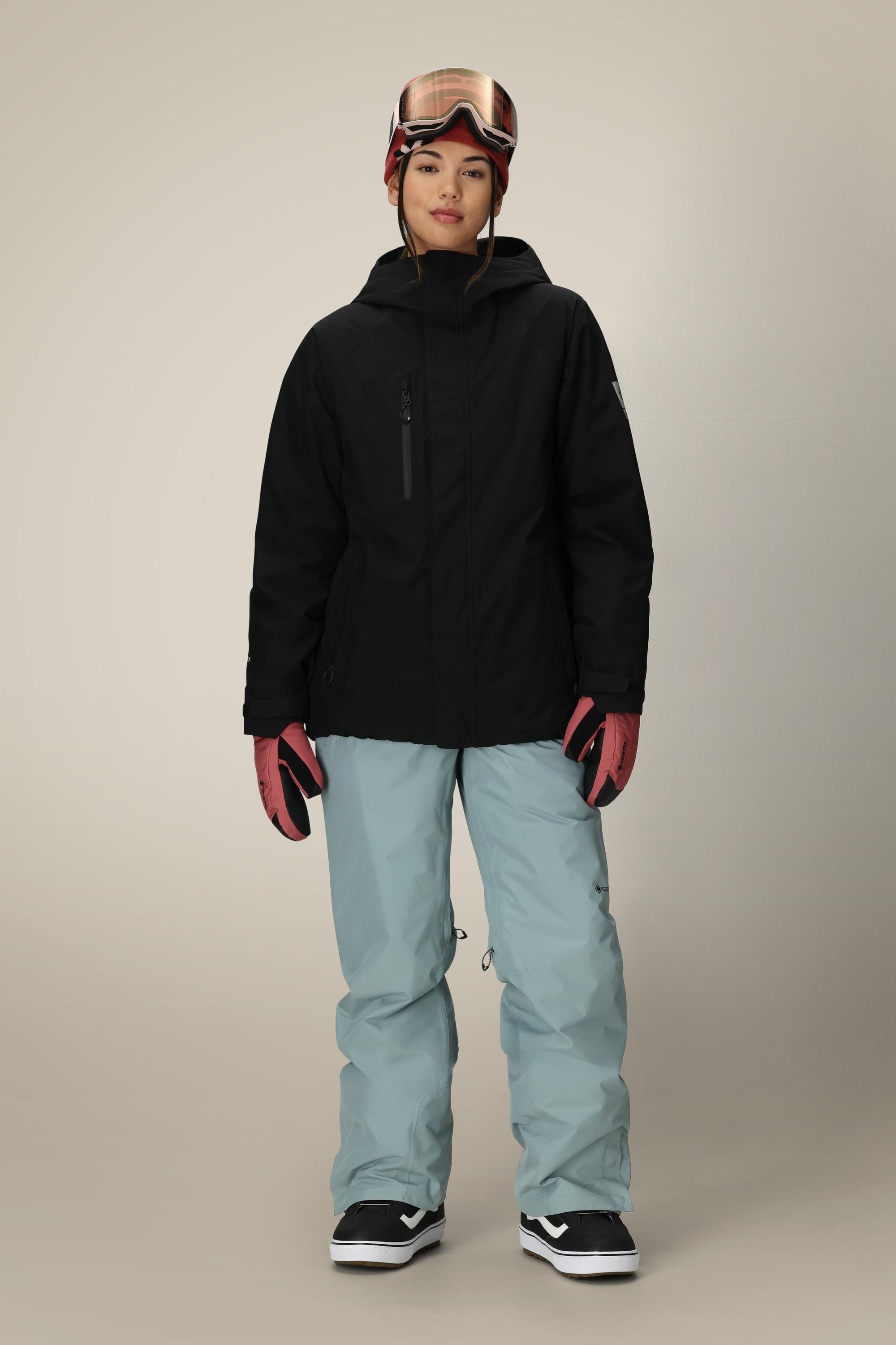A woman stands facing forward in a 686 Women's GORE-TEX Willow™ Insulated Jacket, light blue snow pants, pink gloves, black-and-white snowboard boots, and pink ski goggles against a neutral background.