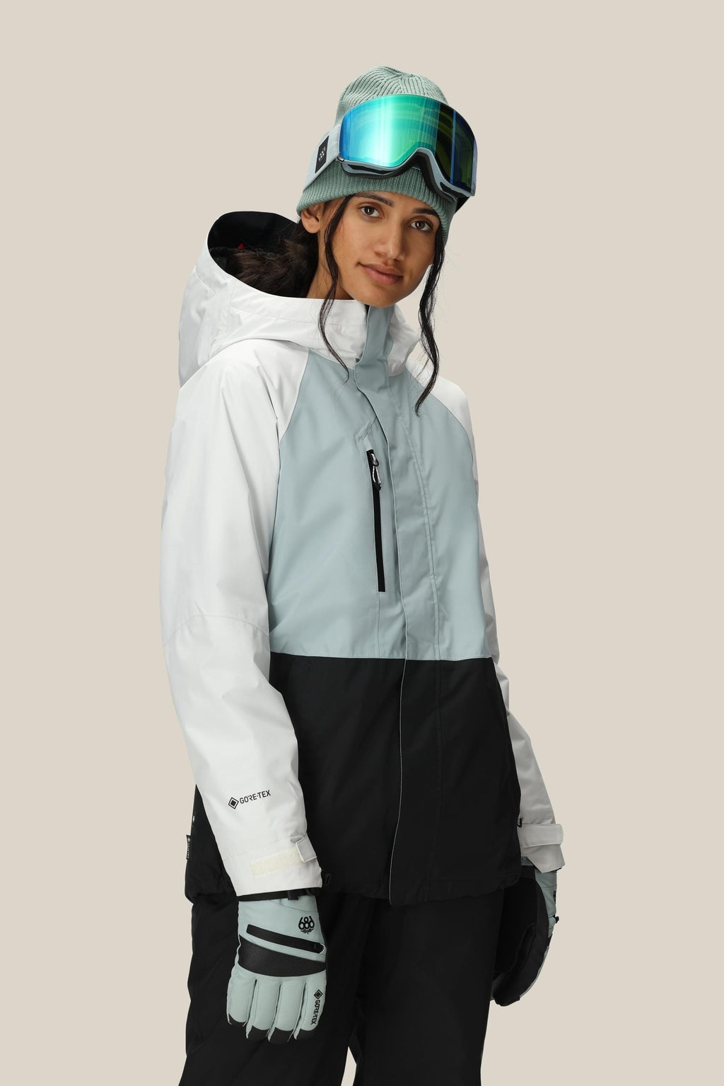 A woman wearing the 686 Women's GORE-TEX Willow™ Insulated Jacket stands against a plain beige background, paired with black snow pants, light blue gloves, a green beanie, and ski goggles.