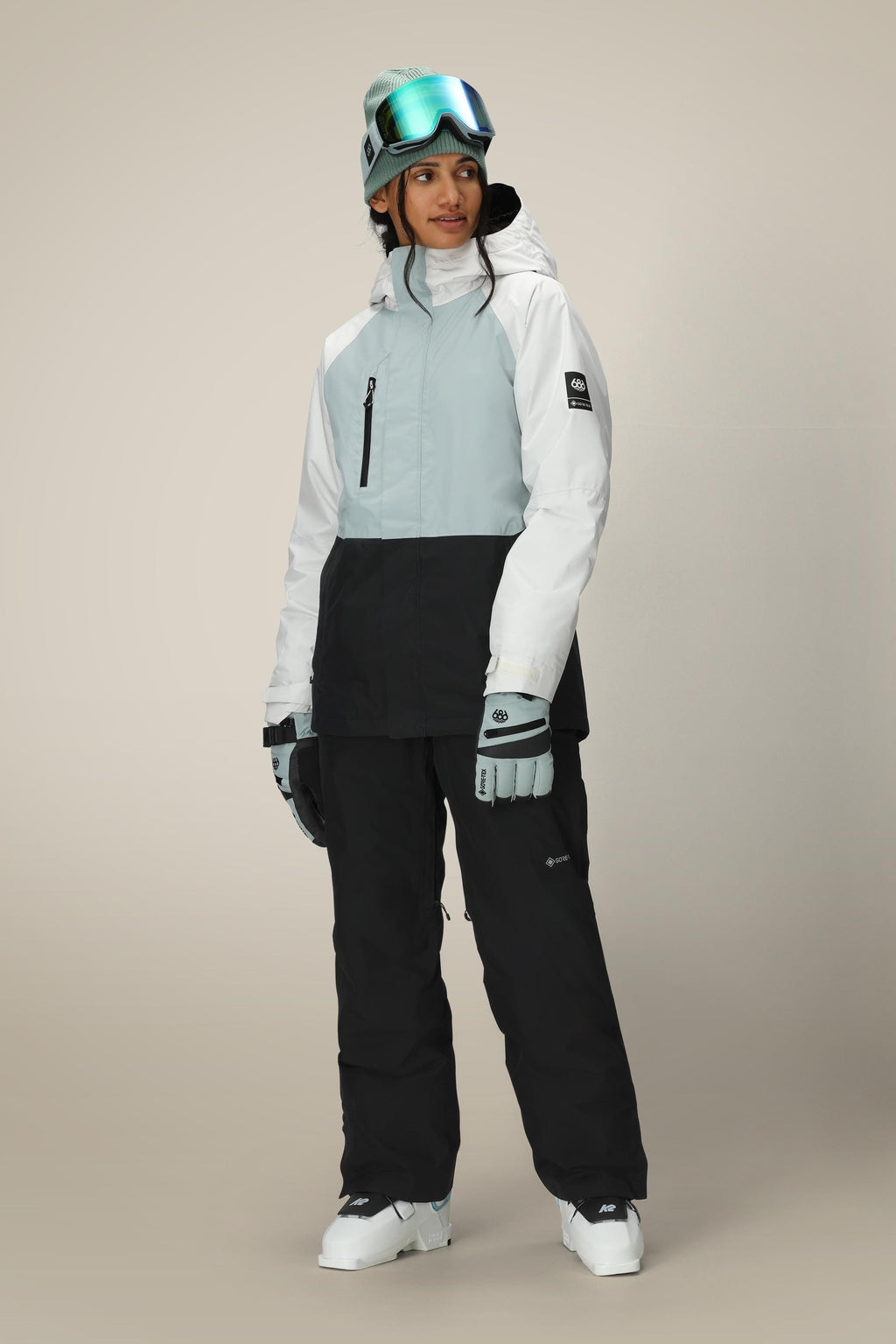 A woman in a 686 Women's GORE-TEX Willow™ Insulated Jacket, black snow pants, gloves, and gray beanie with ski goggles stands against a plain light background.