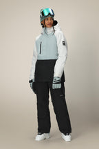 A woman in a 686 Women's GORE-TEX Willow™ Insulated Jacket, black snow pants, gloves, and gray beanie with ski goggles stands against a plain light background.