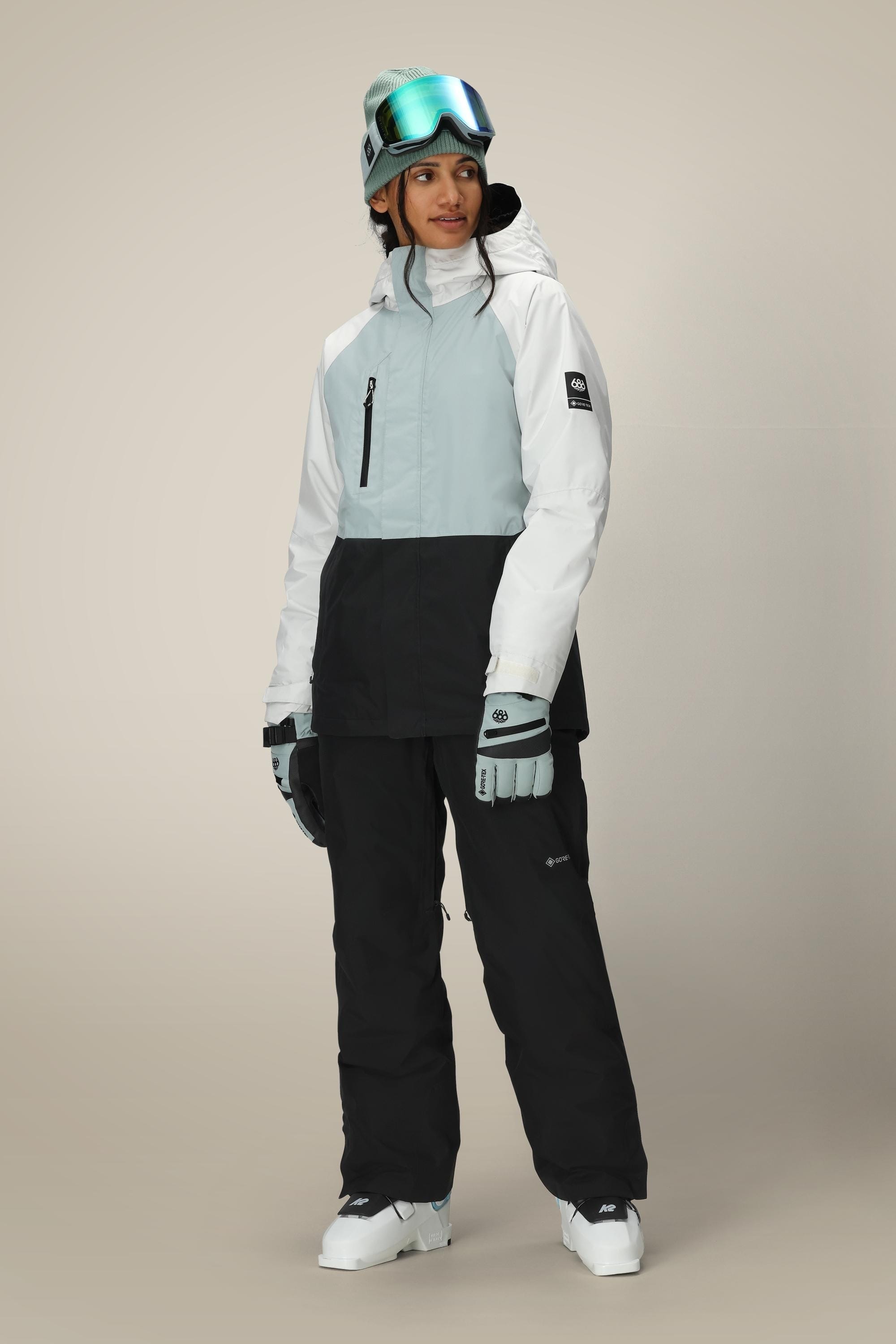 A woman in a 686 Women's GORE-TEX Willow™ Insulated Jacket, black snow pants, gloves, and gray beanie with ski goggles stands against a plain light background.