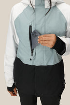 A person wearing the 686 Women's GORE-TEX Willow™ Insulated Jacket in a white, light blue, and black color-block puts a smartphone into its zippered chest pocket. Only their hand and part of the face are visible. Brand: 686.