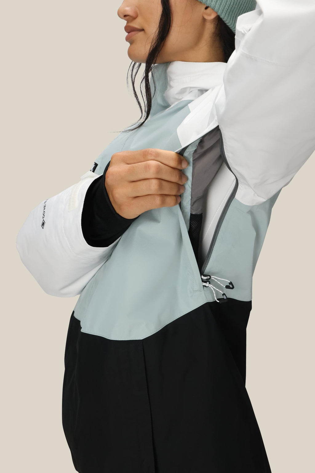 A person wearing the 686 Women's GORE-TEX Willow™ Insulated Jacket with white sleeves, a light blue upper, and a black lower section unzips a side vent. They're also sporting a green beanie.