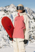 A person in a pink and white 686 Women's GORE-TEX Willow™ Insulated Jacket holds a red snowboard while standing on a snowy mountain with snow-covered peaks in the background.