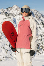 A person in a pink and white 686 Women's GORE-TEX Willow™ Insulated Jacket holds a red snowboard while standing on a snowy mountain with snow-covered peaks in the background.