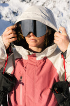 Wearing the 686 Women's GORE-TEX Willow™ Insulated Jacket, a person adjusts their hood and ski goggles against a backdrop of snowy mountains.