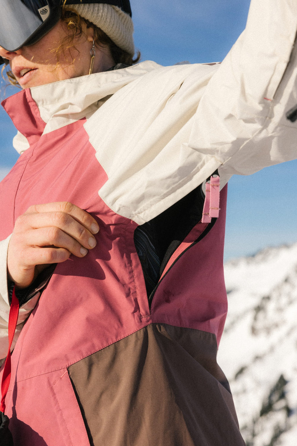 A person wearing the 686 Women's GORE-TEX Willow™ Insulated Jacket in pink, brown, and white opens a zippered vent under their arm, with snow-covered mountains and a blue sky in the background.