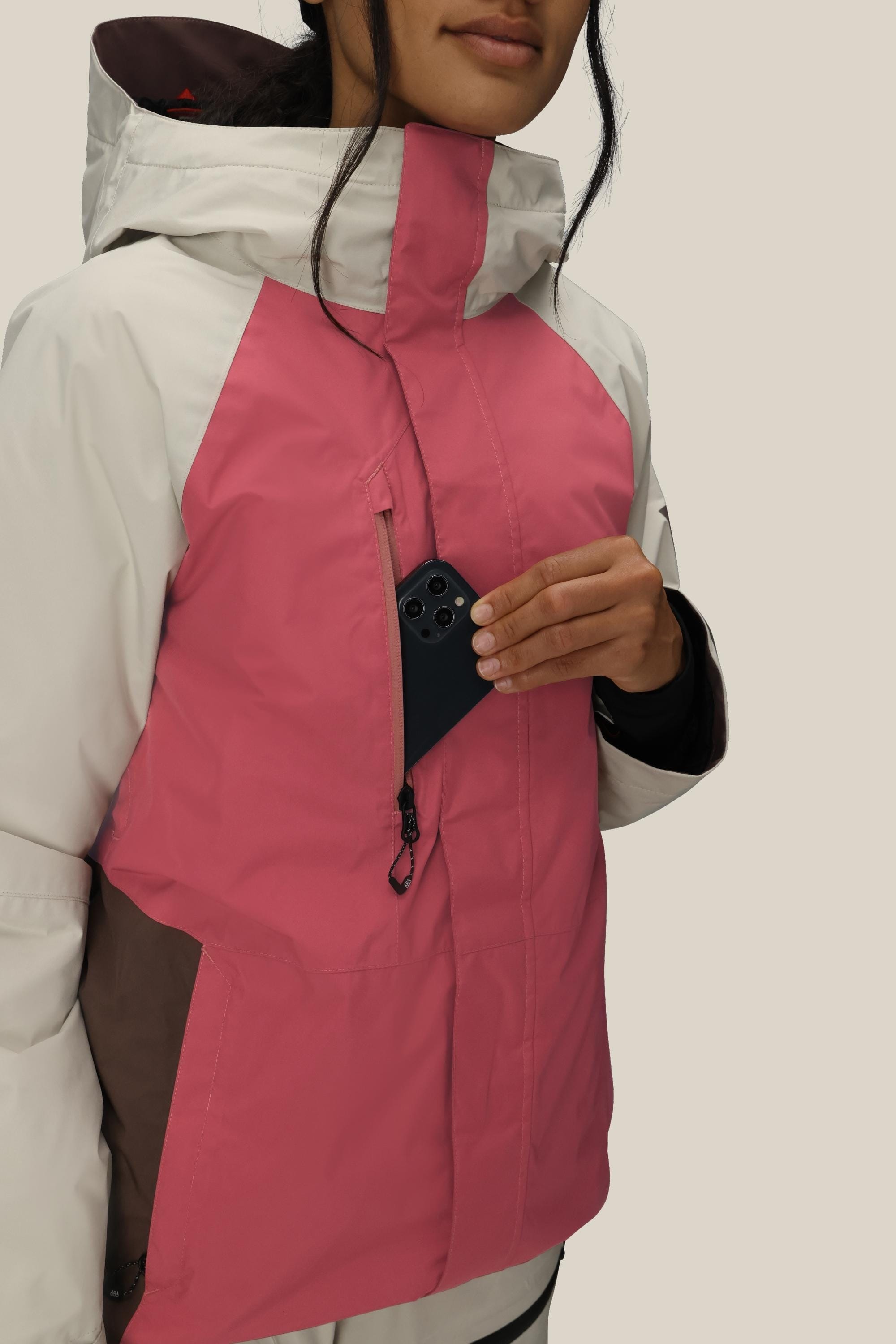 A woman wearing the 686 Women's GORE-TEX Willow™ Insulated Jacket in pink and white puts a black smartphone into its zippered chest pocket. The background is plain and light-colored.