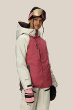 A woman in a 686 Women's GORE-TEX Willow™ Insulated Jacket, white ski pants, pink and white gloves, and pink ski goggles stands against a light background, smiling slightly.