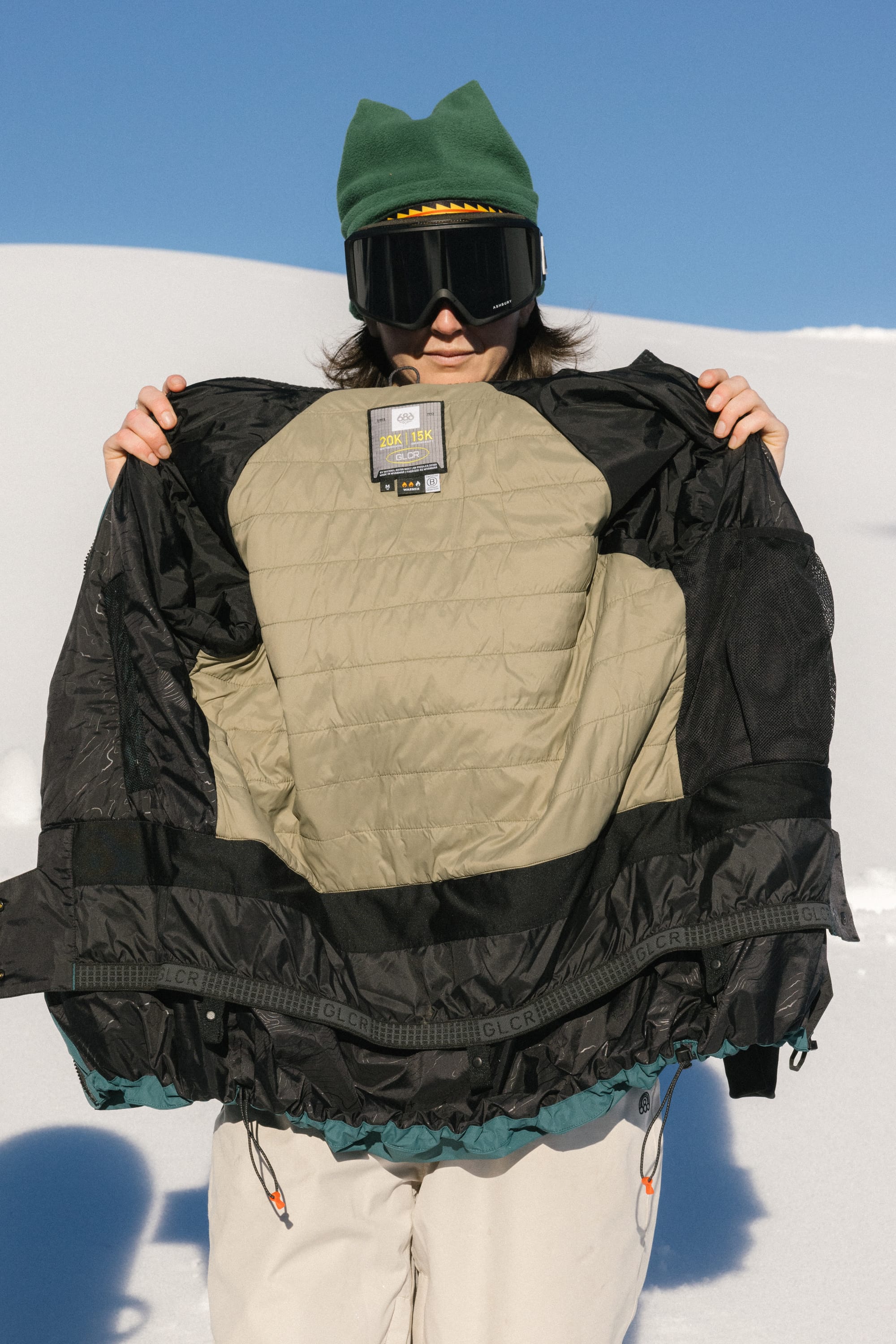 Wearing a green hat and ski goggles, a person stands in the snow on a sunny day, holding open the 686 Women's Whisper™ Insulated Jacket to display its interior lining.