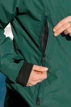 A person wearing the 686 Women's Whisper™ Insulated Jacket in green is unzipping a side pocket, exposing its black mesh lining. Only their hands and part of the 686 jacket with black cuffs are visible.