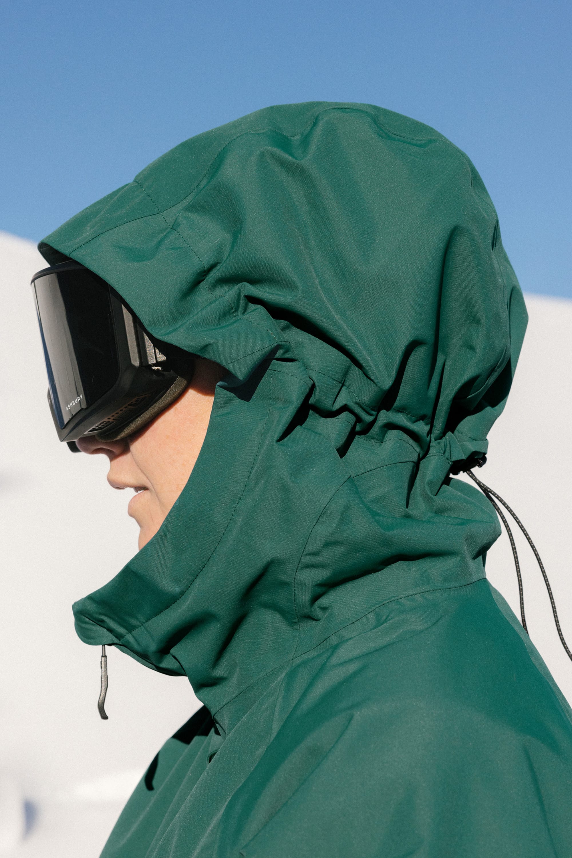 Wearing the 686 Women's Whisper™ Insulated Jacket and black ski goggles, a woman stands outdoors in bright sunlight with a snowy landscape and clear blue sky in the background.