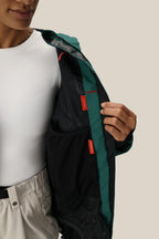 A woman in a white shirt and light pants holds open the green and black inner lining of the 686 Women's Whisper™ Insulated Jacket, revealing multiple interior pockets and red tags.