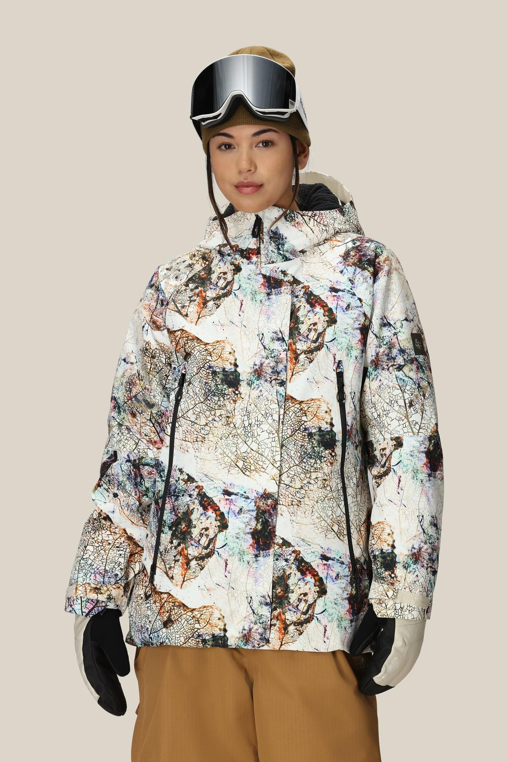 A woman wearing the 686 Women's Whisper™ Insulated Jacket stands against a plain beige background, styled with tan snow pants, black gloves, and ski goggles on her head.