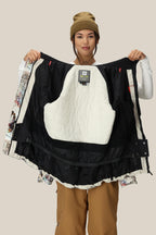 A woman in a tan beanie and brown pants, facing the camera and smiling, holds open the 686 Women's Whisper™ Insulated Jacket to display its white and black quilted interior lining.