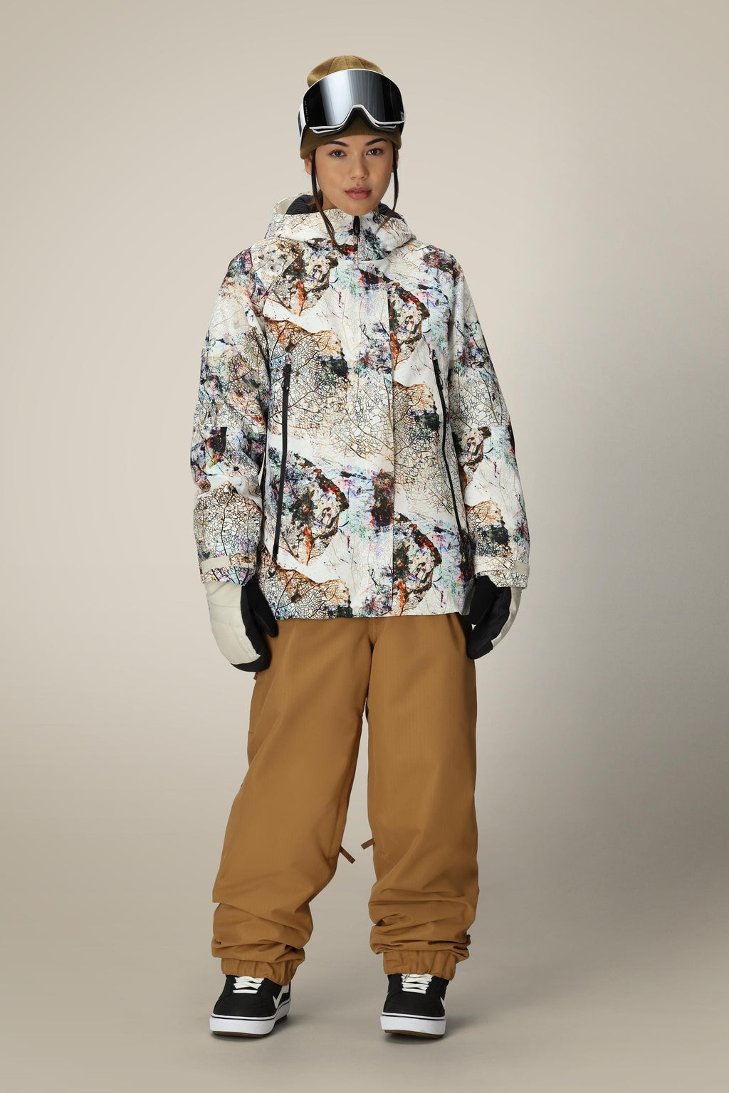 A person wears the 686 Women's Whisper™ Insulated Jacket, tan snow pants, black gloves and snow boots, white ski goggles, and a beige beanie, standing against a plain beige background.