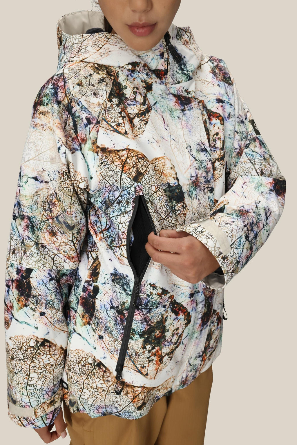 A person in a 686 Women's Whisper™ Insulated Jacket with an abstract pattern and hood unzips a side pocket. Only the lower half of their face is visible against a plain beige background.