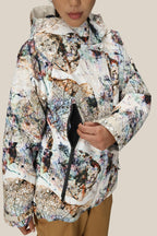 A person in a 686 Women's Whisper™ Insulated Jacket with an abstract pattern and hood unzips a side pocket. Only the lower half of their face is visible against a plain beige background.