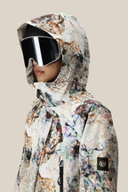 A person wearing the 686 Women's Whisper™ Insulated Jacket—an abstract-patterned, polyester waterproof coat with a hood and 686 logo patch—pairs it with large black ski goggles, standing against a plain beige background.