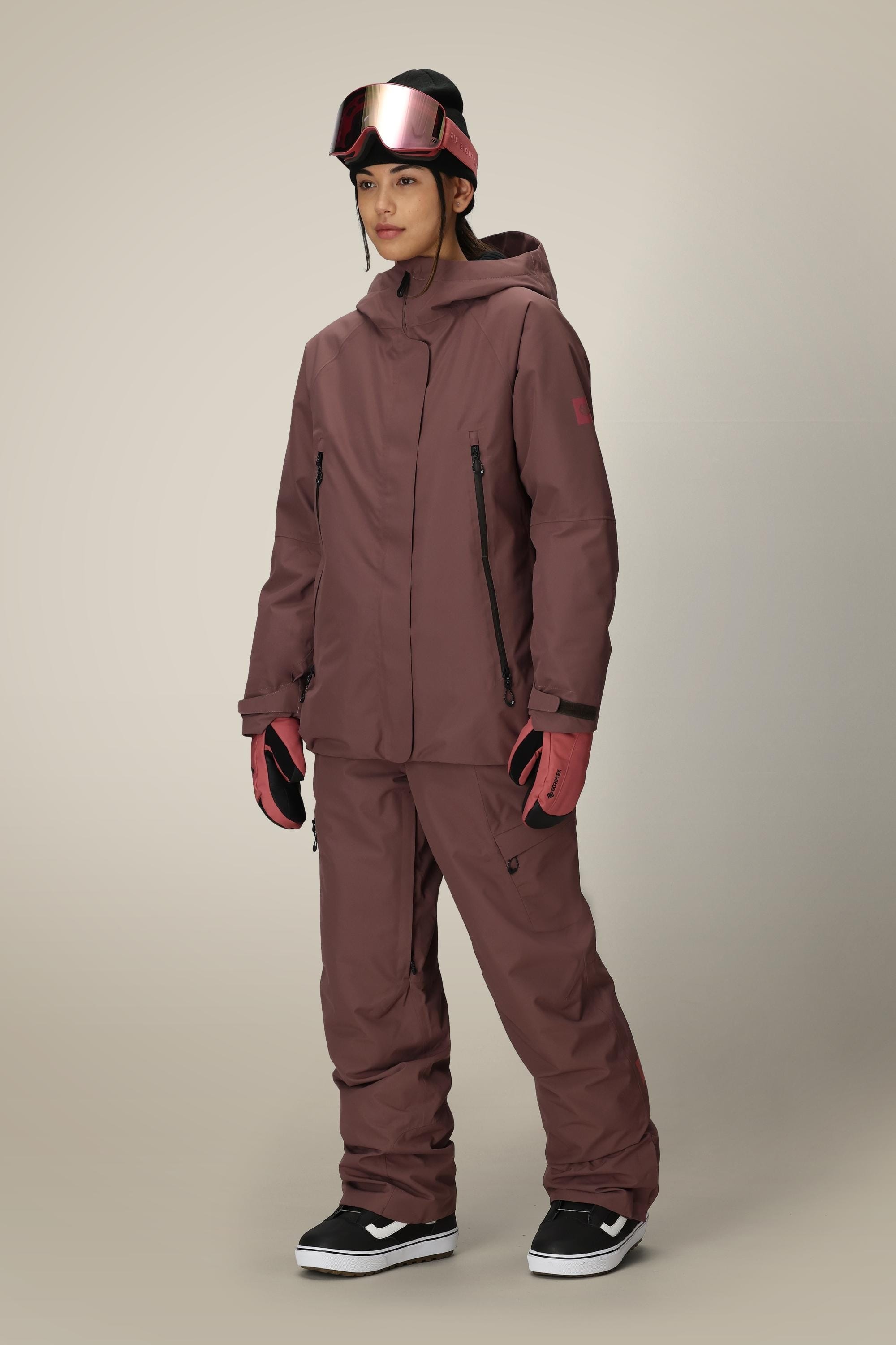 A woman stands against a plain background wearing the 686 Women's Whisper™ Insulated Jacket in maroon, matching pants, pink gloves, a black beanie, and pink ski goggles.