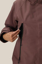 A person in a 686 Women's Whisper™ Insulated Jacket (mauve) unzips a side vertical pocket, showing the black inner lining. Only the upper body and hand are visible.