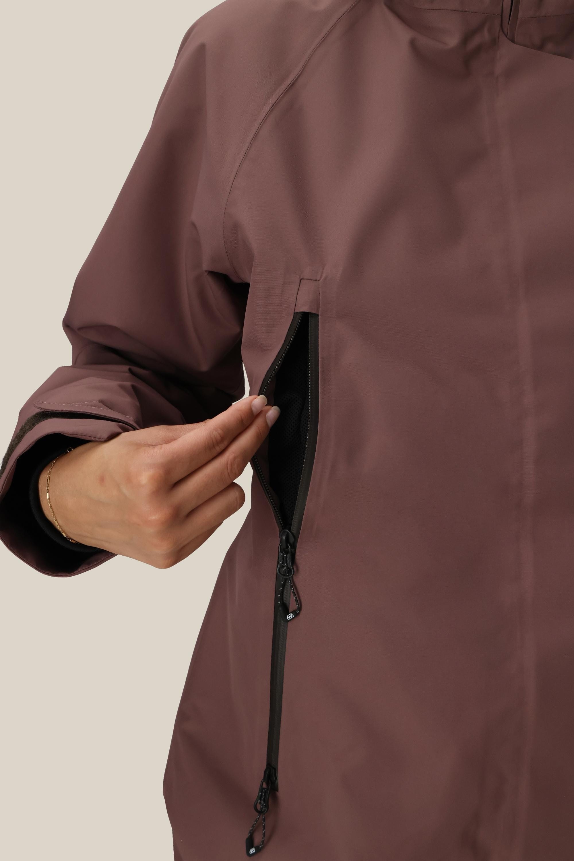 A person in a 686 Women's Whisper™ Insulated Jacket (mauve) unzips a side vertical pocket, showing the black inner lining. Only the upper body and hand are visible.