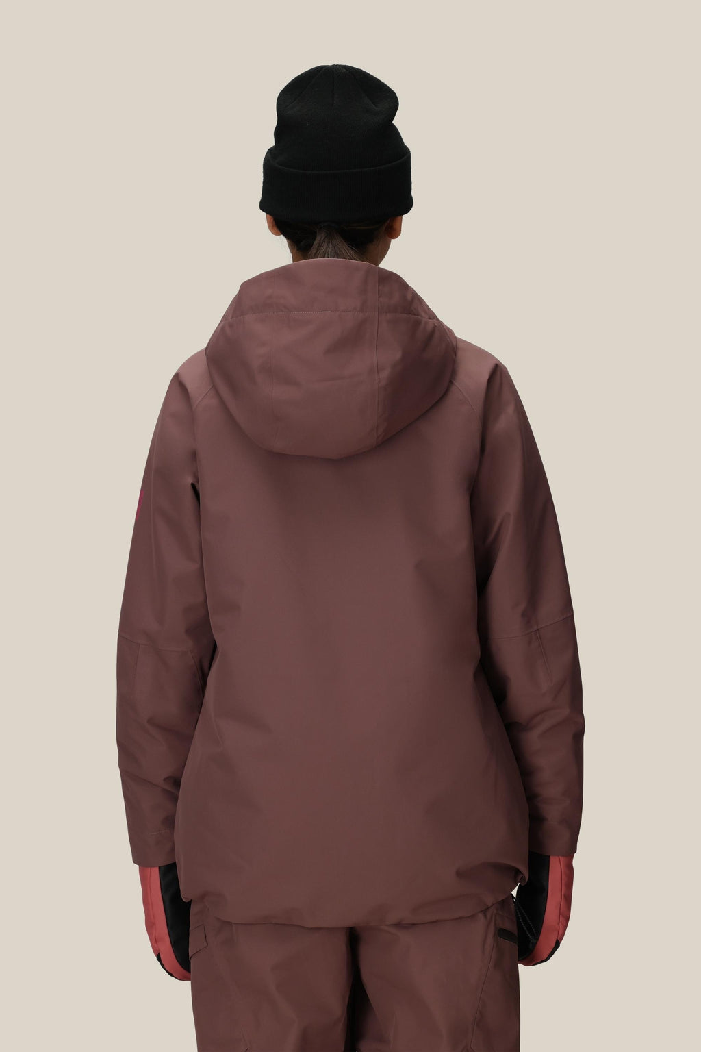 A woman faces away from the camera against a beige background, wearing the 686 Women's Whisper™ Insulated Jacket with matching pants, a black beanie, and pink gloves.